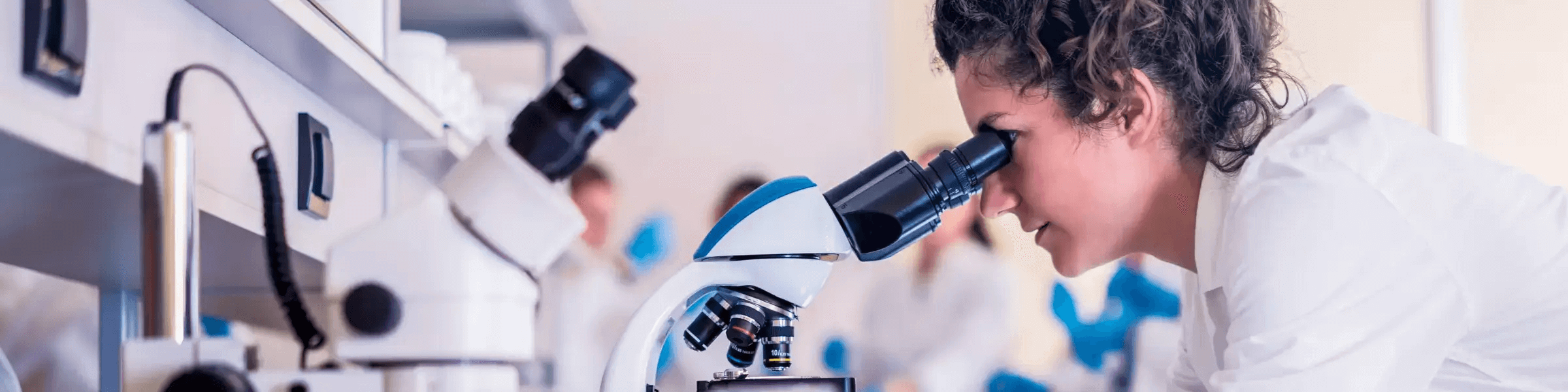 Wissenschaftlerin mit dunklen Haaren und weißem Kittel schaut durch ein Mikroskop, im Hintergrund ist ein Labor zu sehen.