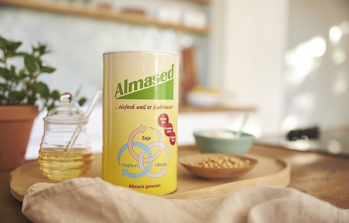 Almased Dose auf einem Tisch mit den drei Hauptzutaten Soja, Joghurt und Honig, die für eine hohe biologische Wertigkeit sorgen.