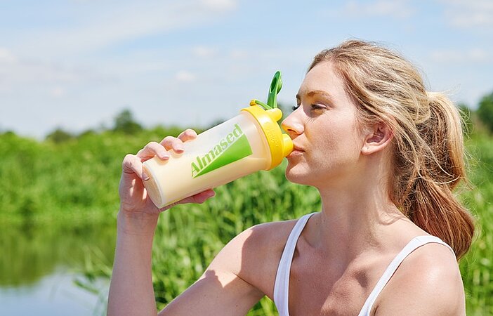 junge-blone-Frau-trinkt-Almased-aus-Shaker-draussen-Natur